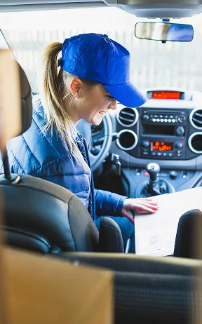A smiling woman with a blue cap and coat sits in a car, surrounded by cardboard boxes, and looks at a document proposing work as a package delivery driver.
