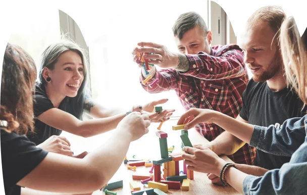 A group of five people enthusiastically builds a tower with colorful wooden blocks on a table, a testament to teamwork and creativity. Bright, natural light streams in.