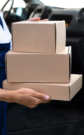 A person in blue overalls is holding three cardboard boxes next to an open delivery van door, indicating a package delivery. The scene exudes efficiency.